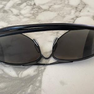 May include: Black sunglasses with dark lenses. The sunglasses have a sleek, modern design with a thin frame and a curved top bar. The lenses are oval-shaped and provide full coverage. The sunglasses are resting on a white and gray marble surface.