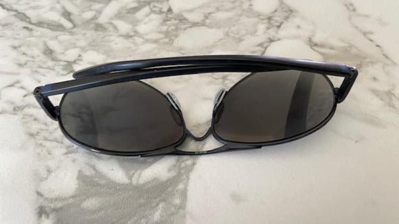 May include: Black sunglasses with dark lenses. The sunglasses have a sleek, modern design with a thin frame and a curved top bar. The lenses are oval-shaped and provide full coverage. The sunglasses are resting on a white and gray marble surface.