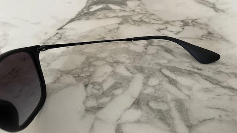 Puede incluir: Gafas de sol negras con acabado mate. Las monturas son cuadradas con lentes degradadas. Las patillas son finas y se curvan en la oreja. Las gafas de sol est&aacute;n sobre una superficie de m&aacute;rmol.