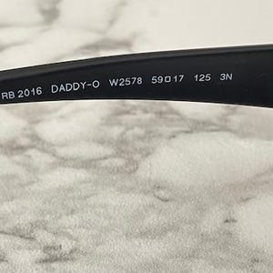 May include: The interior of a matte black sunglass temple arm featuring white printed text that reads RB 2016 DADDY-O W2578 59 17 125 3N. The arm is constructed from a solid black material and includes a visible hinge mechanism on the right side.