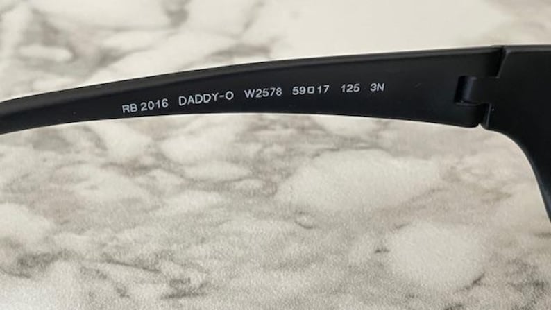 May include: The interior of a matte black sunglass temple arm featuring white printed text that reads RB 2016 DADDY-O W2578 59 17 125 3N. The arm is constructed from a solid black material and includes a visible hinge mechanism on the right side.