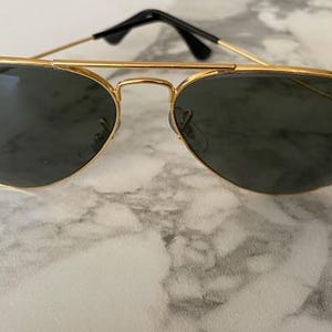 May include: Gold-framed aviator sunglasses with dark lenses. The sunglasses have a double bridge and black temples. The Ray-Ban logo is visible on the left lens. A classic eyewear accessory.