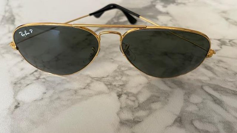 May include: Gold-framed aviator sunglasses with dark lenses. The sunglasses have a double bridge and black temples. The Ray-Ban logo is visible on the left lens. A classic eyewear accessory.