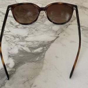 May include: Brown tortoiseshell sunglasses with dark lenses. The sunglasses have a classic, rounded shape and are set against a marble background. The arms of the sunglasses are thin and have a tortoiseshell pattern.