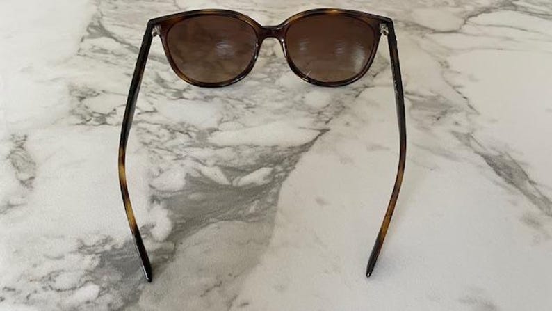 May include: Brown tortoiseshell sunglasses with dark lenses. The sunglasses have a classic, rounded shape and are set against a marble background. The arms of the sunglasses are thin and have a tortoiseshell pattern.