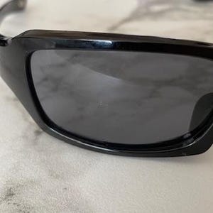 K&ouml;nnte beinhalten: Nahaufnahme einer schwarzen Sonnenbrille mit dunkelgrauen Gl&auml;sern. Die Sonnenbrille hat ein schlankes, modernes Design mit gl&auml;nzendem Finish. Der Rahmen ist dick und umschlie&szlig;t die Gl&auml;ser und bietet einen sportlichen Look. Die Gl&auml;ser sind get&ouml;nt.