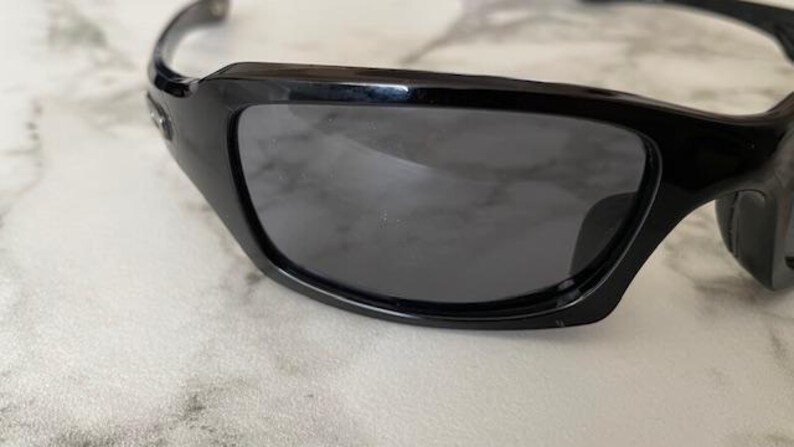 K&ouml;nnte beinhalten: Nahaufnahme einer schwarzen Sonnenbrille mit dunkelgrauen Gl&auml;sern. Die Sonnenbrille hat ein schlankes, modernes Design mit gl&auml;nzendem Finish. Der Rahmen ist dick und umschlie&szlig;t die Gl&auml;ser und bietet einen sportlichen Look. Die Gl&auml;ser sind get&ouml;nt.