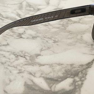 May include: Dark gray sunglasses with a rectangular frame and tinted lenses. The temple arm features the text "OO9102-X558 57 18 137". The sunglasses are set against a marble-patterned background.