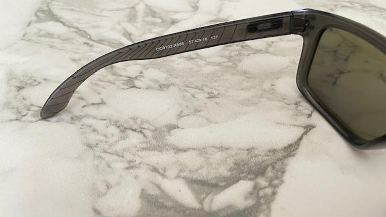 May include: Dark gray sunglasses with a rectangular frame and tinted lenses. The temple arm features the text "OO9102-X558 57 18 137". The sunglasses are set against a marble-patterned background.