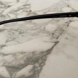 May include: Black sunglasses with a rounded lens shape and a thin, black frame. The temple arm has the text "D00973-39". The lenses are dark, and the sunglasses are set against a marble-like surface.