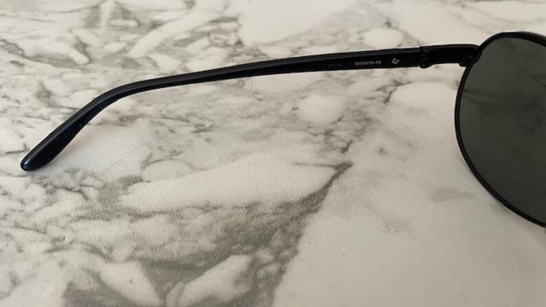 May include: Black sunglasses with a rounded lens shape and a thin, black frame. The temple arm has the text "D00973-39". The lenses are dark, and the sunglasses are set against a marble-like surface.