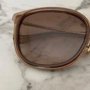 May include: Close-up of sunglasses with brown lenses and a light brown frame. The sunglasses have a rounded shape and a silver metal bridge. The hinges are gold-colored. The lenses have a gradient tint.