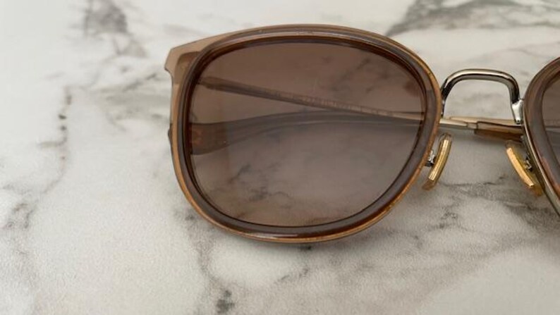 May include: Close-up of sunglasses with brown lenses and a light brown frame. The sunglasses have a rounded shape and a silver metal bridge. The hinges are gold-colored. The lenses have a gradient tint.