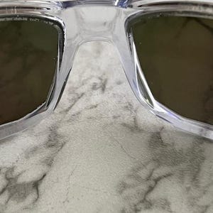 May include: Close-up of clear-framed sunglasses with dark lenses. The sunglasses have a classic rectangular shape and a transparent frame. The lenses appear to be a dark shade, providing contrast against the clear frame. The image is a close-up, highlighting the details of the eyewear.