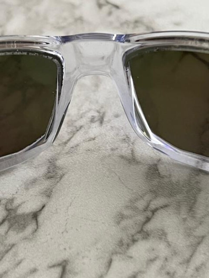 May include: Close-up of clear-framed sunglasses with dark lenses. The sunglasses have a classic rectangular shape and a transparent frame. The lenses appear to be a dark shade, providing contrast against the clear frame. The image is a close-up, highlighting the details of the eyewear.