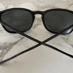 Puede incluir: Gafas de sol negras con lentes oscuras. Las gafas tienen un dise&ntilde;o cl&aacute;sico con montura redonda y patillas rectas. Las patillas est&aacute;n cruzadas en la imagen. Las gafas descansan sobre una superficie de m&aacute;rmol blanco y gris.