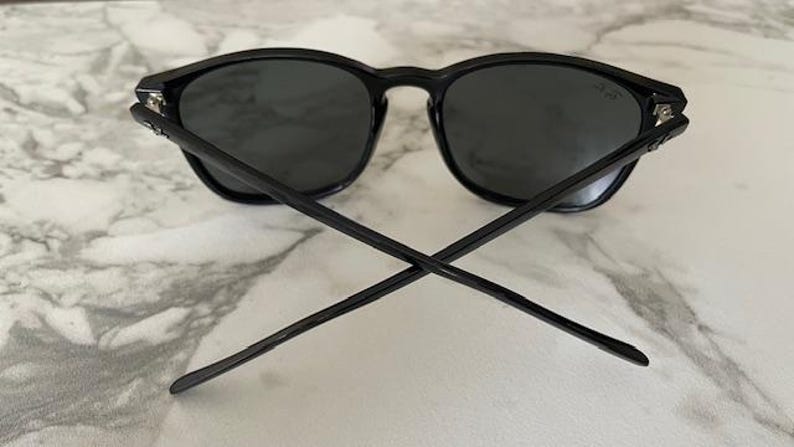 Puede incluir: Gafas de sol negras con lentes oscuras. Las gafas tienen un dise&ntilde;o cl&aacute;sico con montura redonda y patillas rectas. Las patillas est&aacute;n cruzadas en la imagen. Las gafas descansan sobre una superficie de m&aacute;rmol blanco y gris.