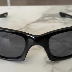 K&ouml;nnte beinhalten: Schwarze Sonnenbrille mit einem schlanken, modernen Design. Die Sonnenbrille hat eine gl&auml;nzende Oberfl&auml;che und dunkle Gl&auml;ser. Die Rahmen sind dick und umschlie&szlig;en das Gesicht und bieten einen sportlichen Look. Die Sonnenbrille liegt auf einer hellen Oberfl&auml;che.