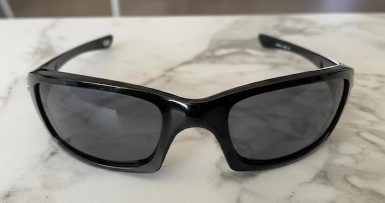 K&ouml;nnte beinhalten: Schwarze Sonnenbrille mit einem schlanken, modernen Design. Die Sonnenbrille hat eine gl&auml;nzende Oberfl&auml;che und dunkle Gl&auml;ser. Die Rahmen sind dick und umschlie&szlig;en das Gesicht und bieten einen sportlichen Look. Die Sonnenbrille liegt auf einer hellen Oberfl&auml;che.