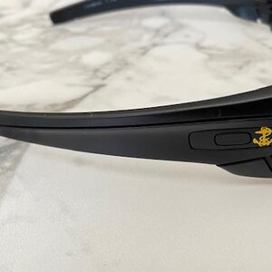 May include: Black sunglasses with a gold anchor emblem on the temple. The sunglasses have a sleek, modern design with dark lenses and a curved frame. The glasses are set against a white and grey marble background.