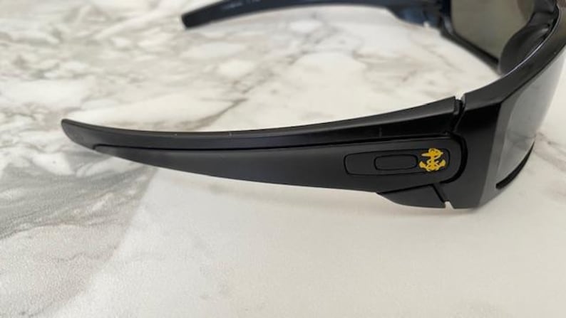 May include: Black sunglasses with a gold anchor emblem on the temple. The sunglasses have a sleek, modern design with dark lenses and a curved frame. The glasses are set against a white and grey marble background.
