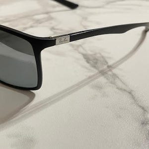 May include: Black sunglasses with gray lenses. The sunglasses have a rectangular frame and a silver logo on the temple. The sunglasses are on a white marble surface.