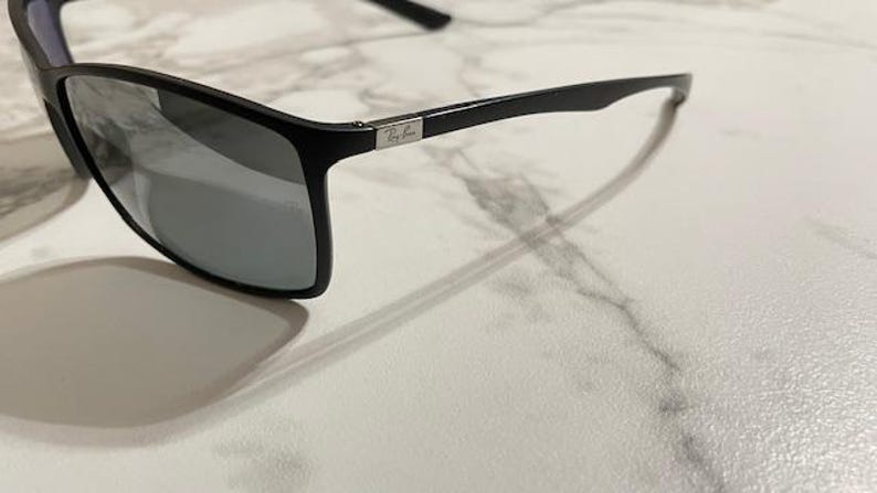 May include: Black sunglasses with gray lenses. The sunglasses have a rectangular frame and a silver logo on the temple. The sunglasses are on a white marble surface.