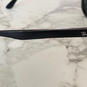 Puede incluir: Gafas de sol negras con el logotipo de Ray-Ban en la patilla. Las gafas tienen un acabado brillante y est&aacute;n sobre una superficie de m&aacute;rmol. Las lentes son oscuras y la montura es gruesa y rectangular.