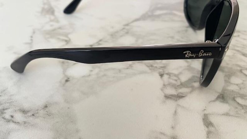 Puede incluir: Gafas de sol negras con el logotipo de Ray-Ban en la patilla. Las gafas tienen un acabado brillante y est&aacute;n sobre una superficie de m&aacute;rmol. Las lentes son oscuras y la montura es gruesa y rectangular.