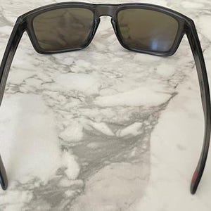 May include: Dark gray sunglasses with rectangular lenses and a matching frame. The sunglasses are resting on a white and gray marble surface. The temples have a slight curve.