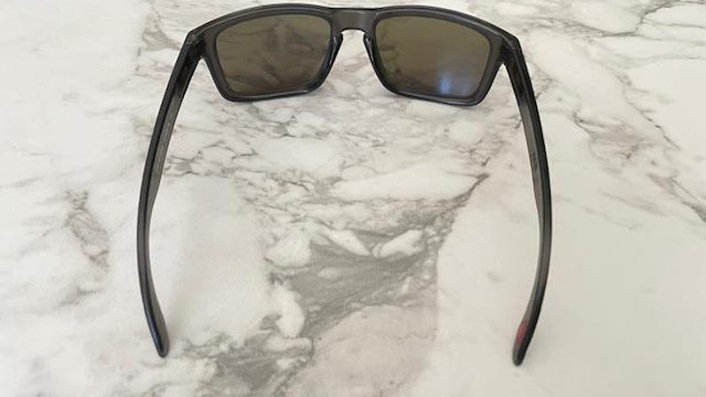 May include: Dark gray sunglasses with rectangular lenses and a matching frame. The sunglasses are resting on a white and gray marble surface. The temples have a slight curve.