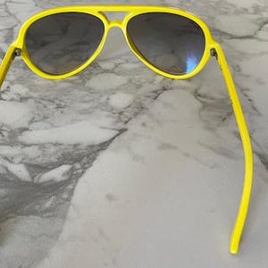 May include: Bright yellow aviator sunglasses with dark lenses. The sunglasses have a classic double bridge and a sleek, modern design. The frames are a vibrant yellow, offering a bold and stylish look. These sunglasses are perfect for sunny days and outdoor activities.