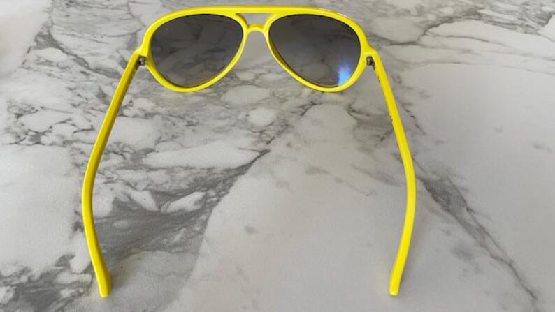 May include: Bright yellow aviator sunglasses with dark lenses. The sunglasses have a classic double bridge and a sleek, modern design. The frames are a vibrant yellow, offering a bold and stylish look. These sunglasses are perfect for sunny days and outdoor activities.