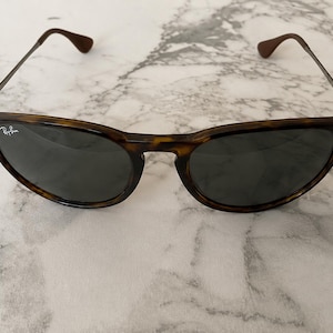 K&ouml;nnte beinhalten: Sonnenbrille mit Schildpattmuster und dunklen Gl&auml;sern. Die Fassungen sind braun und schwarz, mit d&uuml;nnen Metallb&uuml;geln und braunen Ohrenteilen. Das Ray-Ban-Logo ist auf dem linken Glas sichtbar.