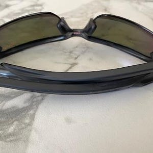 Peut inclure: Lunettes de soleil noires avec une monture incurv&eacute;e et des verres teint&eacute;s. Les lunettes sont pli&eacute;es, mettant en valeur le design &eacute;pur&eacute; et les charni&egrave;res. Les verres ont une teinte d&eacute;grad&eacute;e, passant d'une teinte plus fonc&eacute;e en haut &agrave; une plus claire en bas.