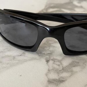K&ouml;nnte beinhalten: Schwarze Sonnenbrille mit rechteckigem Rahmen und dunklen Gl&auml;sern. Die Sonnenbrille hat ein schlankes, modernes Design. Der Rahmen besteht aus gl&auml;nzendem schwarzem Material. Die Gl&auml;ser sind dunkelgrau.