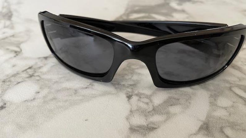 K&ouml;nnte beinhalten: Schwarze Sonnenbrille mit rechteckigem Rahmen und dunklen Gl&auml;sern. Die Sonnenbrille hat ein schlankes, modernes Design. Der Rahmen besteht aus gl&auml;nzendem schwarzem Material. Die Gl&auml;ser sind dunkelgrau.