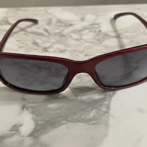 May include: A pair of burgundy sunglasses with dark gray lenses. The sunglasses have a rectangular shape with a slightly curved top frame and temples. The glasses are resting on a white and gray marble surface.