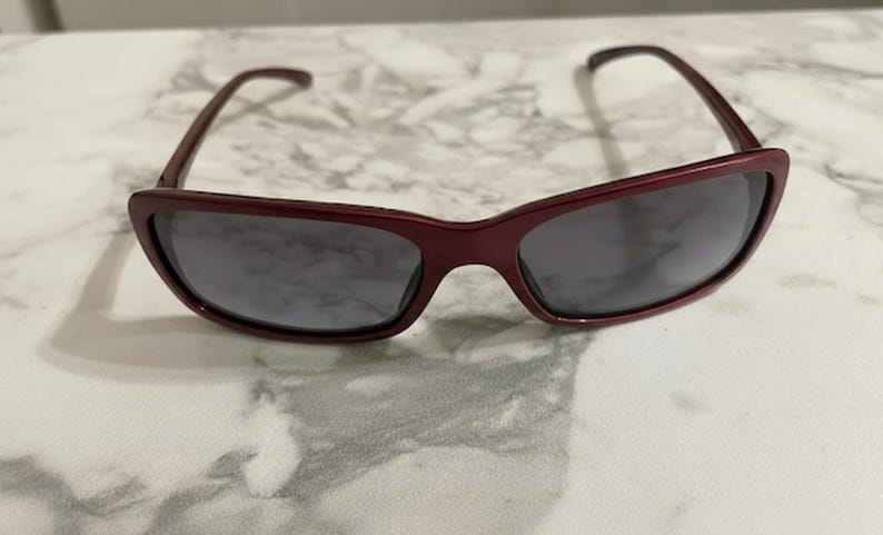 May include: A pair of burgundy sunglasses with dark gray lenses. The sunglasses have a rectangular shape with a slightly curved top frame and temples. The glasses are resting on a white and gray marble surface.