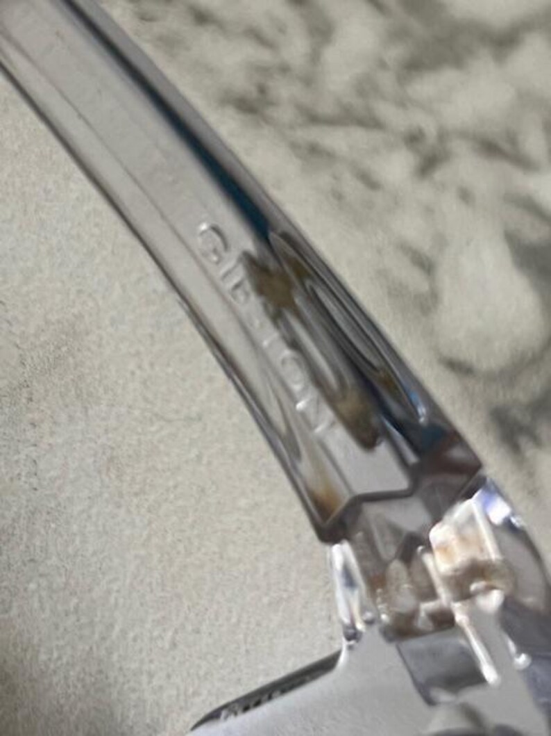 May include: Close-up of a clear plastic object with the word "GIBSON" embossed on the side. The object has a curved shape and appears to be part of a larger item. The background is a blurred, light-colored surface.