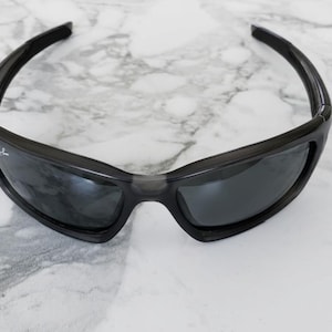 May include: Black sunglasses with a wraparound design. The sunglasses have dark lenses and a black frame. The Ray-Ban logo is visible on the left lens. The sunglasses are on a white and gray marble surface.