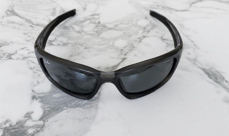 May include: Black sunglasses with a wraparound design. The sunglasses have dark lenses and a black frame. The Ray-Ban logo is visible on the left lens. The sunglasses are on a white and gray marble surface.