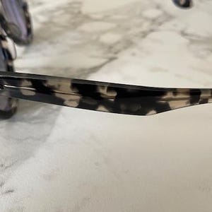 Puede incluir: Primer plano de unas gafas de sol con un estampado moteado en blanco y negro. El logotipo de Ray-Ban es visible en la patilla. Las lentes son de color oscuro.