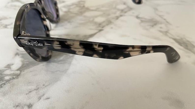 Puede incluir: Primer plano de unas gafas de sol con un estampado moteado en blanco y negro. El logotipo de Ray-Ban es visible en la patilla. Las lentes son de color oscuro.
