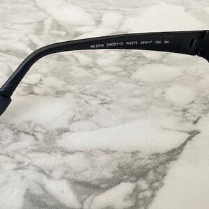 May include: Black sunglasses featuring a matte frame and dark lenses. The inner temple arm displays the white printed text RB 2018 DADDY-O W2578 59017 125 3N. The temple arm curves downward at the end and includes a flexible black rubberized tip.