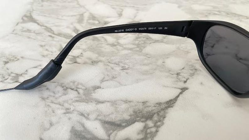 May include: Black sunglasses featuring a matte frame and dark lenses. The inner temple arm displays the white printed text RB 2018 DADDY-O W2578 59017 125 3N. The temple arm curves downward at the end and includes a flexible black rubberized tip.