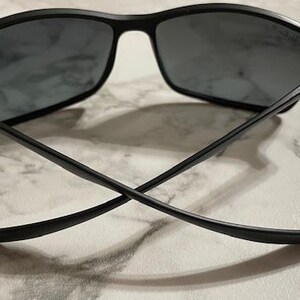 May include: Black sunglasses with a rectangular frame and dark lenses. The temples are thin and cross each other. The sunglasses are set against a light-colored, marbled surface.