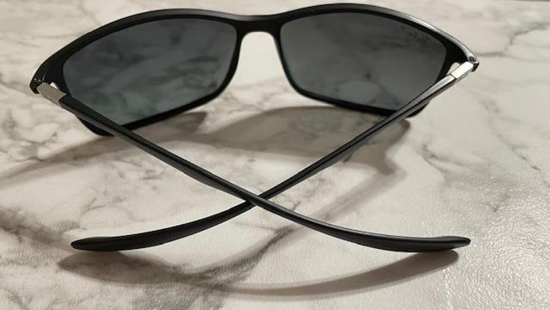 May include: Black sunglasses with a rectangular frame and dark lenses. The temples are thin and cross each other. The sunglasses are set against a light-colored, marbled surface.