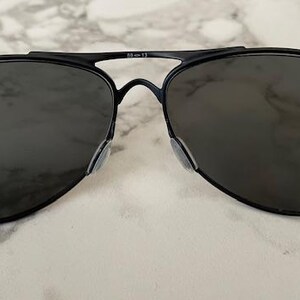 May include: Black aviator sunglasses with dark lenses. The sunglasses have a thin, black metal frame and nose pads. The lenses are a dark gray color, and the overall design is sleek and modern.