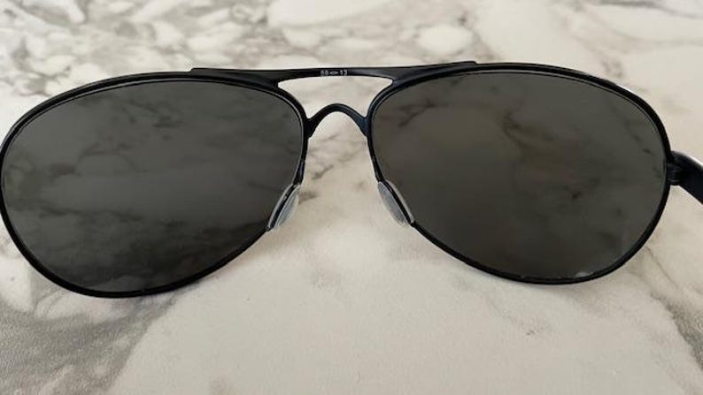 May include: Black aviator sunglasses with dark lenses. The sunglasses have a thin, black metal frame and nose pads. The lenses are a dark gray color, and the overall design is sleek and modern.